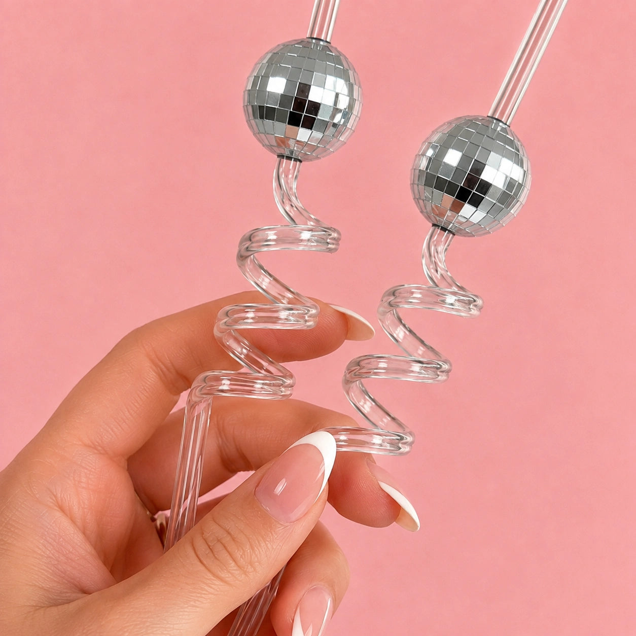 A hand with a French manicure holds two clear spiral straws, each topped with a silver disco ball, against a solid pink background.