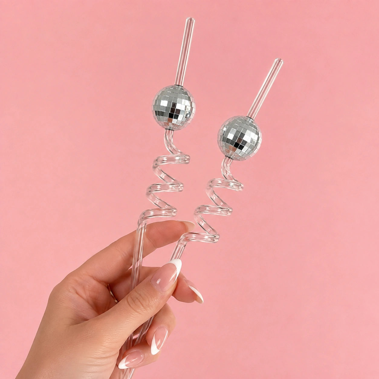 Two clear glass spiral straws with mirrored disco ball tops, held in a hand against a solid pink background.