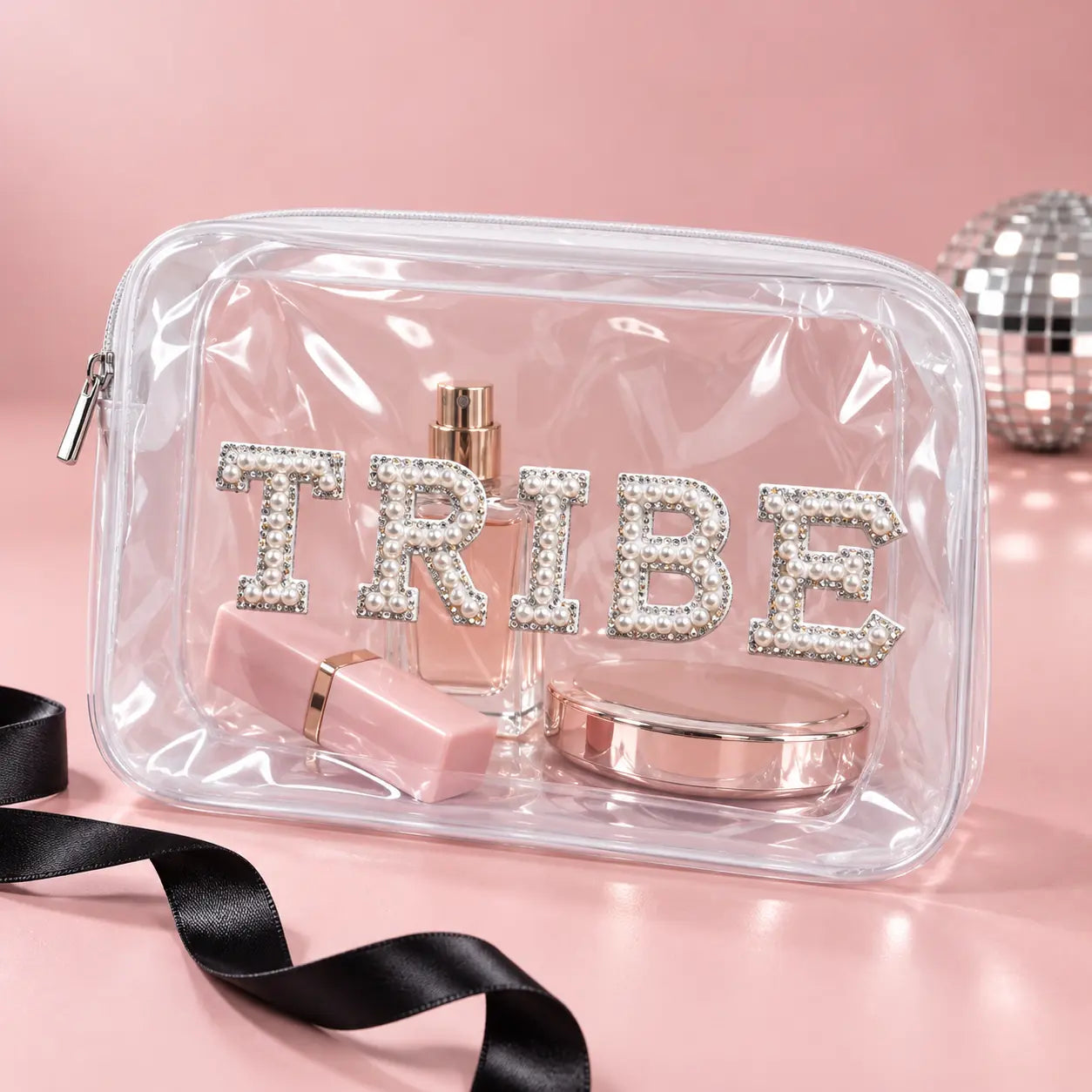 A clear zip cosmetic pouch with the word "TRIBE" spelled out in large rhinestone letters, shown on a pink surface with makeup items inside and a black ribbon in the foreground.