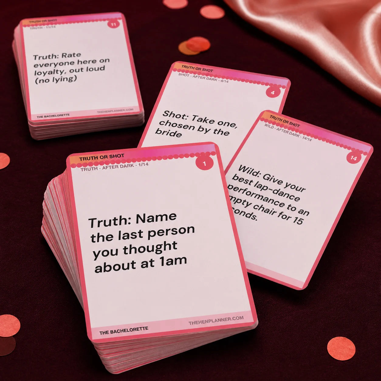 A stack of pink and white bachelorette party truth or dare cards arranged on a dark red surface with scattered confetti and a satin fabric backdrop. 