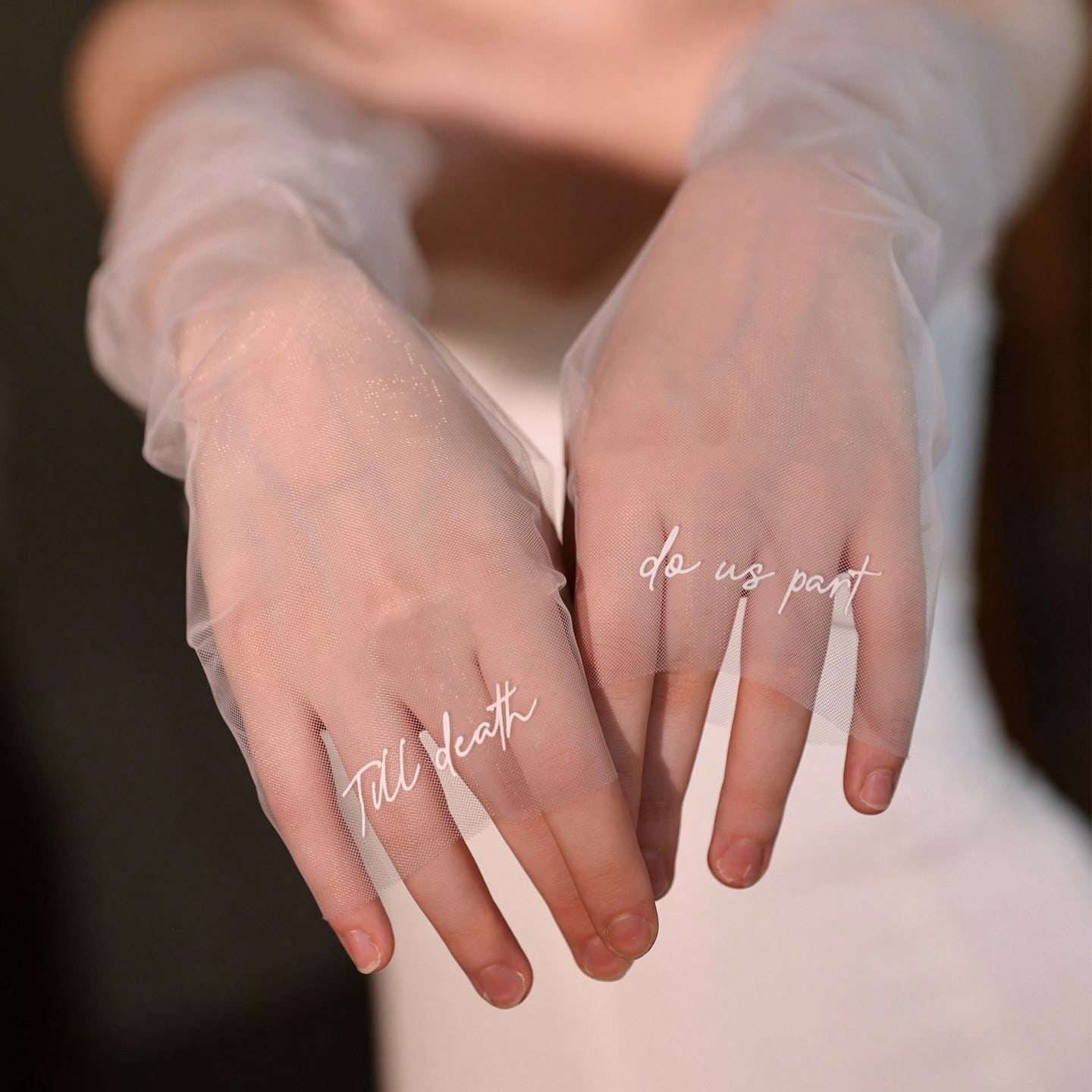 A close-up of a person wearing sheer, semi-transparent long gloves over their hands and wrists, with white script text overlays reading 'do us part' and 'Till death'. The background is a soft, out-of-focus neutral tone.
