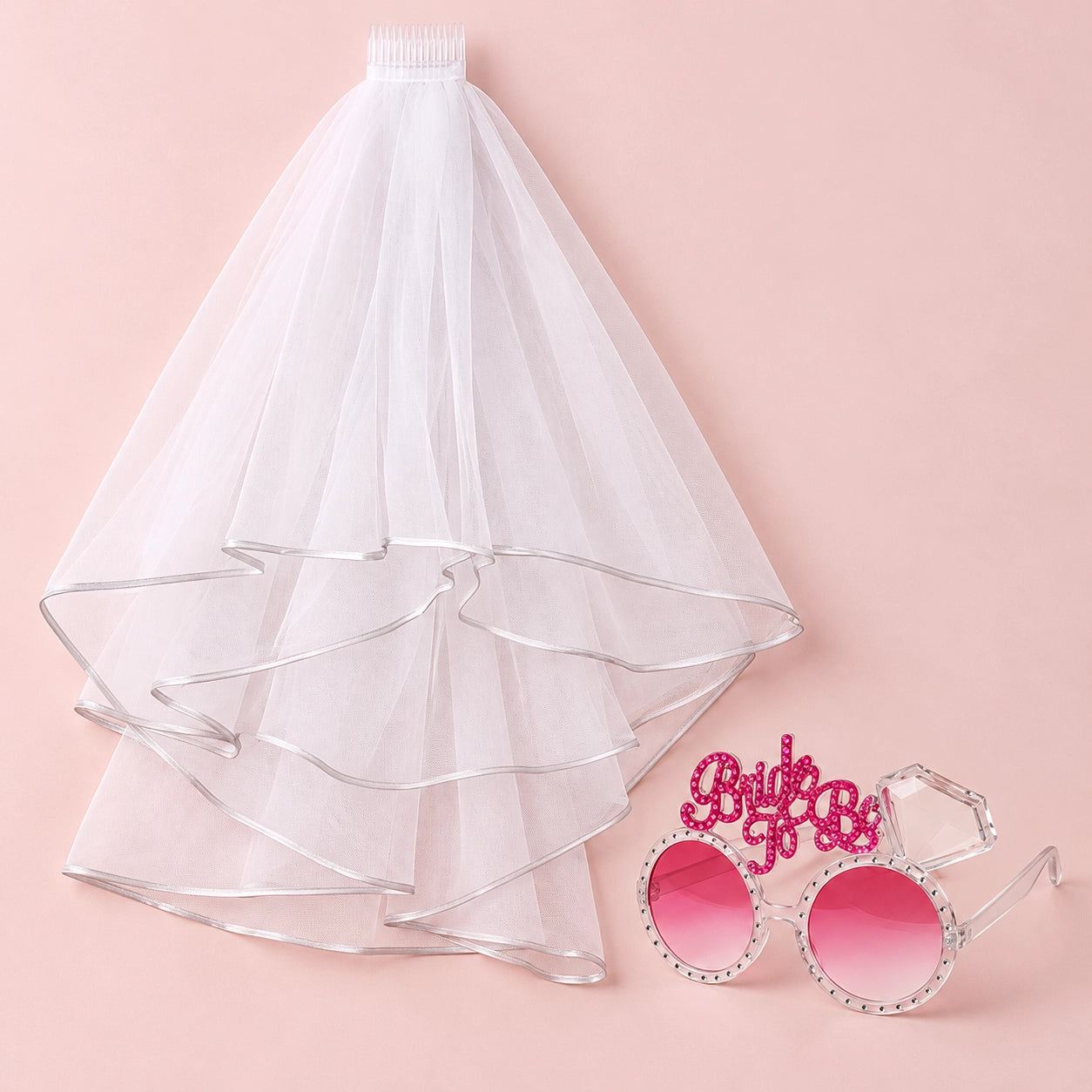 A white bridal veil with layered ruffles and a matching white headband, displayed next to novelty round sunglasses with pink lenses and a pink rhinestone 'Bride to Be' headband on a solid pink background.