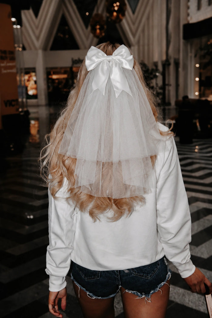 Person wearing a white jumper with a sheer overlay and large bow, standing in an indoor setting.