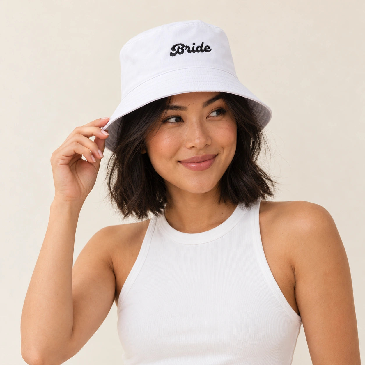 A woman wearing a white bucket hat with the word 'Bride' embroidered in black script on the front, paired with a white ribbed tank top against a plain beige background.