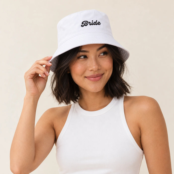 A woman wearing a white bucket hat with the word 'Bride' embroidered in black script on the front, paired with a white ribbed tank top against a plain beige background.