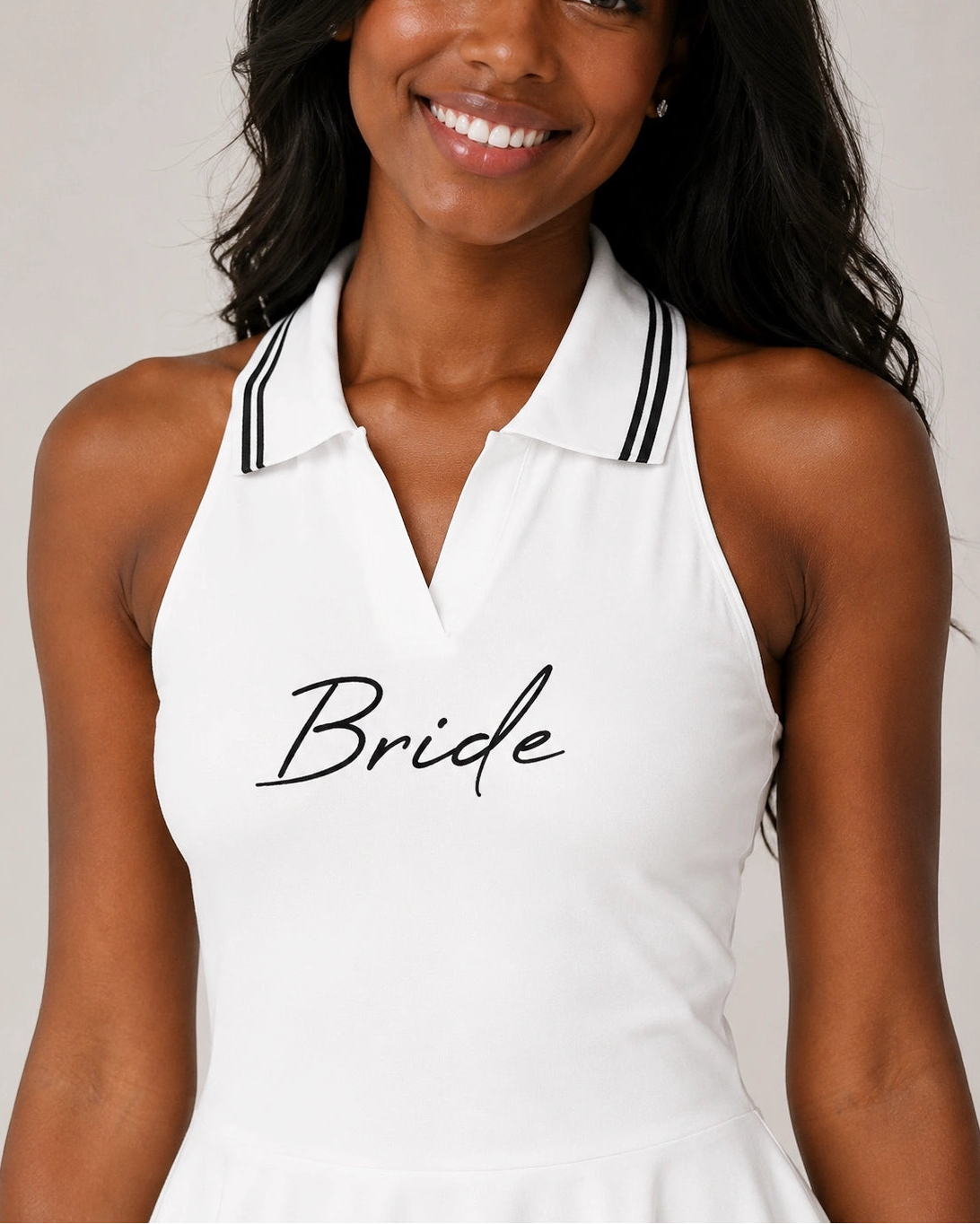 A smiling woman wearing a white halter-neck dress with a polo-style collar trimmed in black stripes and the word 'Bride' printed in black script across the chest, against a light gray studio background.