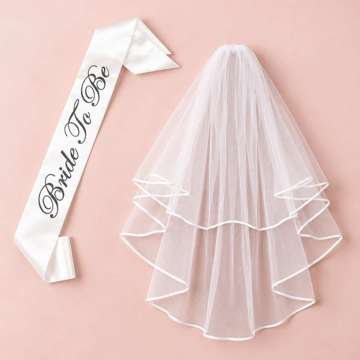White satin Bride To Be sash with dark script beside a two-tier white veil on a blush pink background.