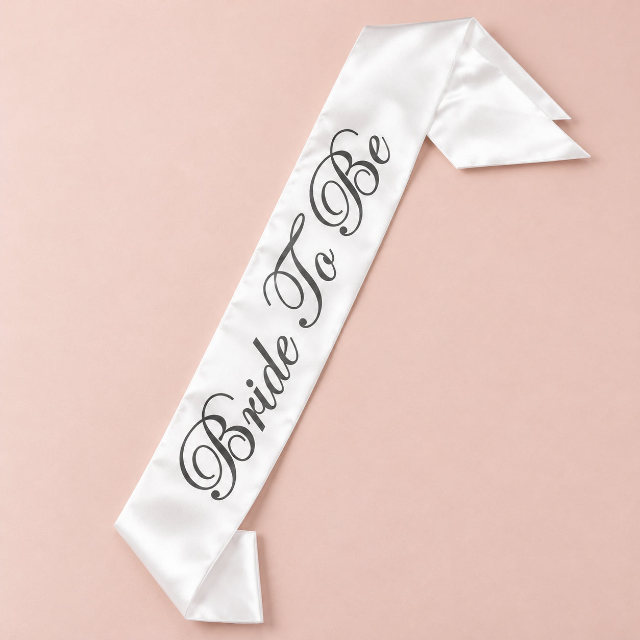 White satin Bride To Be sash with dark calligraphy, arranged diagonally on a blush pink background.