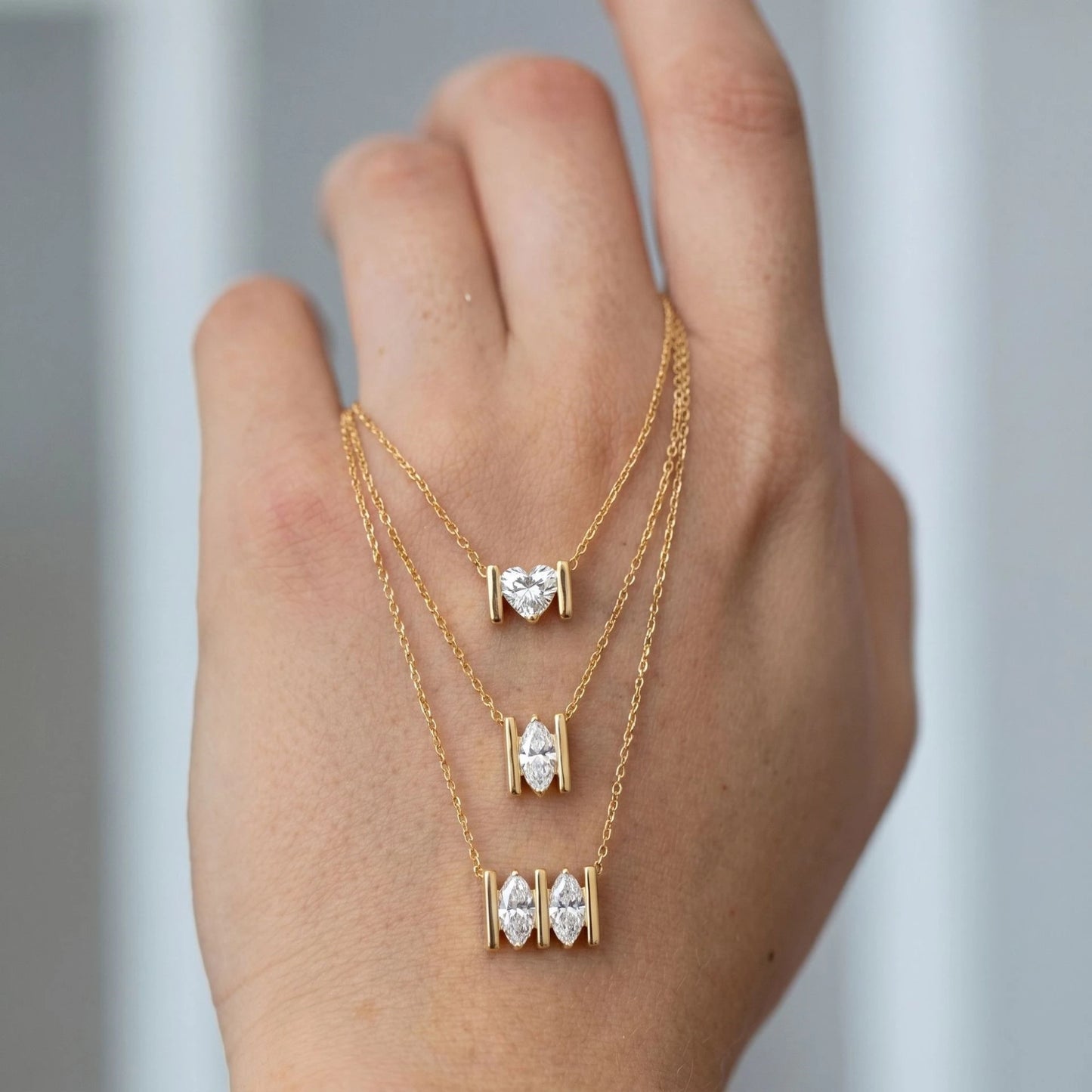 A close-up of a hand wearing three layered gold necklaces with clear marquise-cut gemstones, set against a soft, blurred light gray background.
