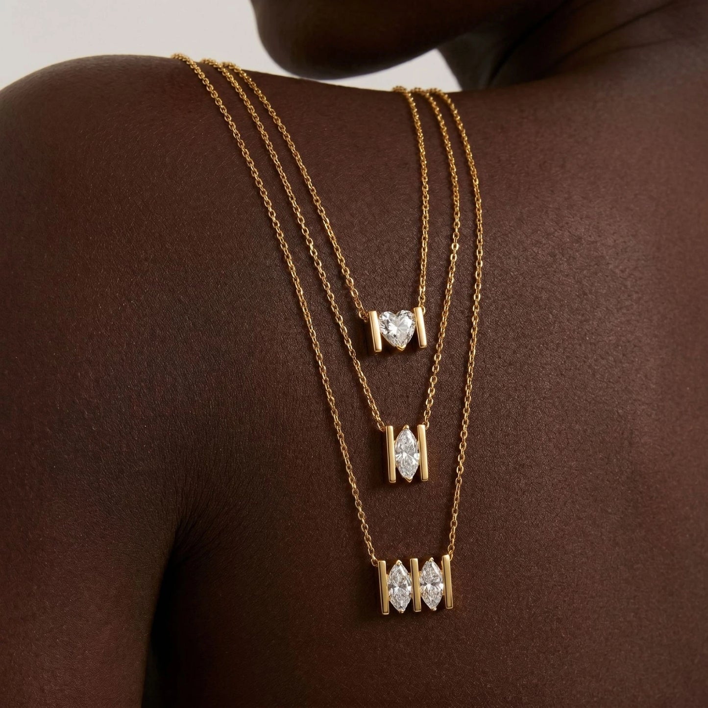 Three layered gold necklaces with heart-shaped and marquise-cut stone pendants, displayed on a model's dark brown shoulder.