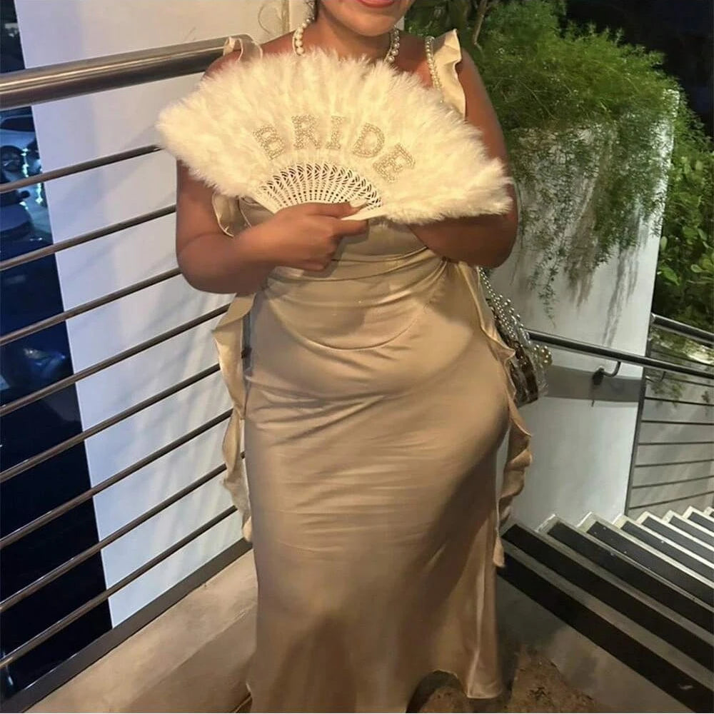 Woman in a beige dress holding a white fan with 'Bride' written on it, standing outdoors.