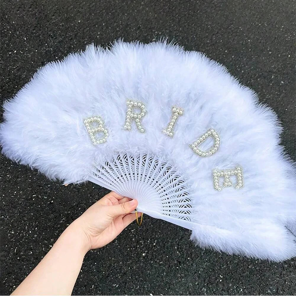 Light blue feather fan with 'Bride' in rhinestones held by a hand on a dark surface