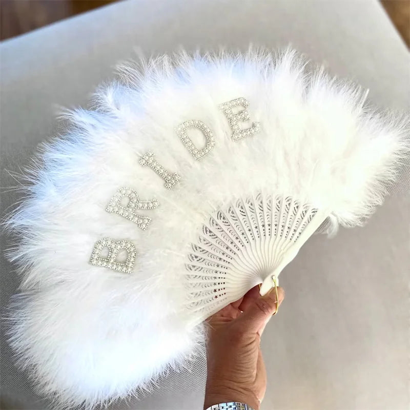 White feathered hand fan with 'BRIDE' embellishments held by a hand on a light background