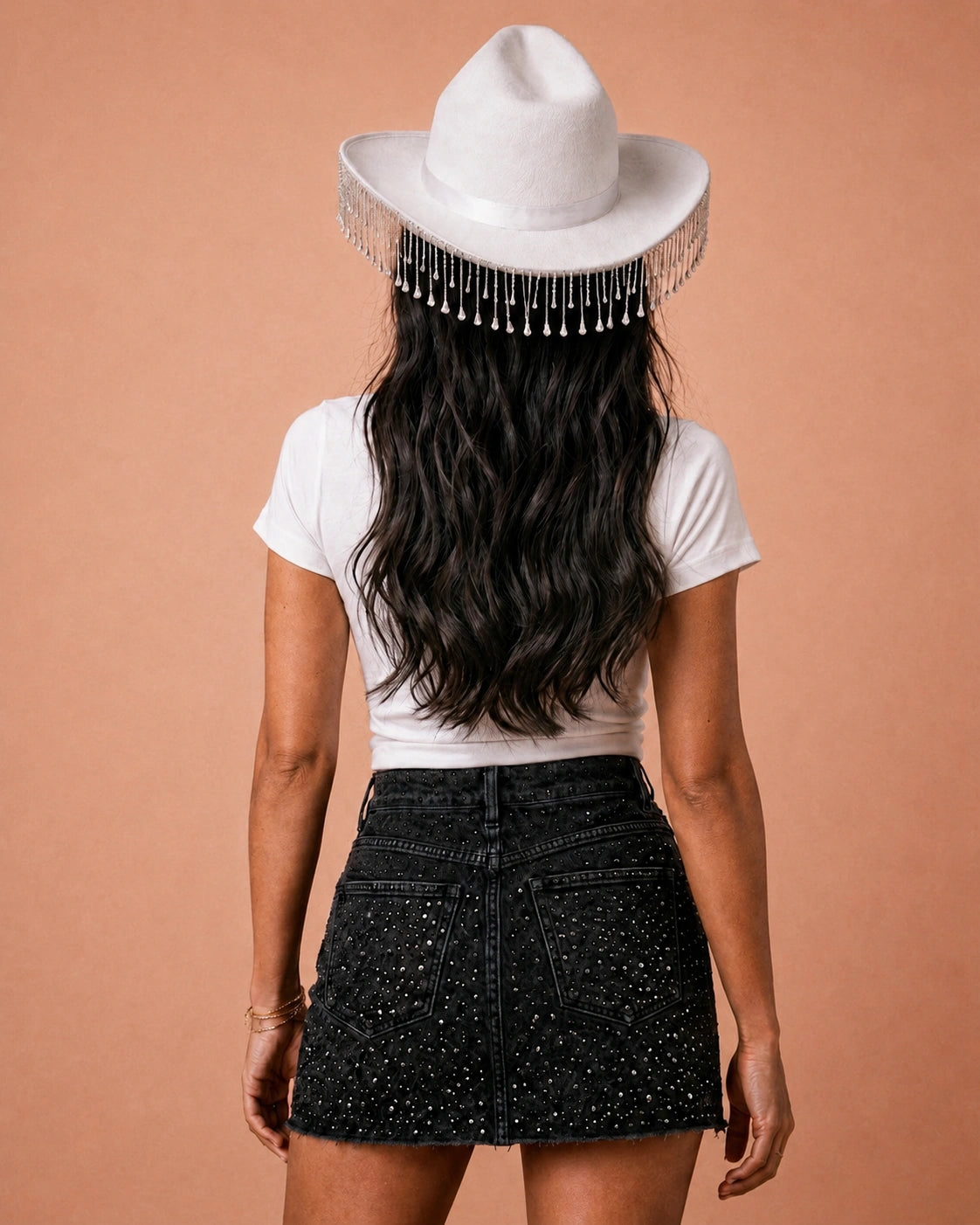 Person wearing a white t-shirt, black embellished skirt, and white cowgirl hat with fringe on a beige background