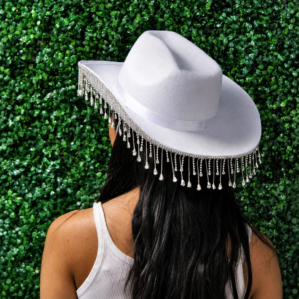 Person wearing a white cowboy hat with fringe against a green hedge background
