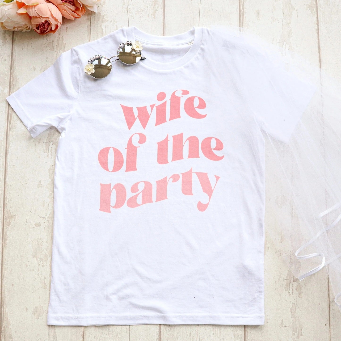 Flat lay of a white “wife of the party” hen party t-shirt with pale pink text, styled with bridal sunglasses and a white veil on a light wooden background.