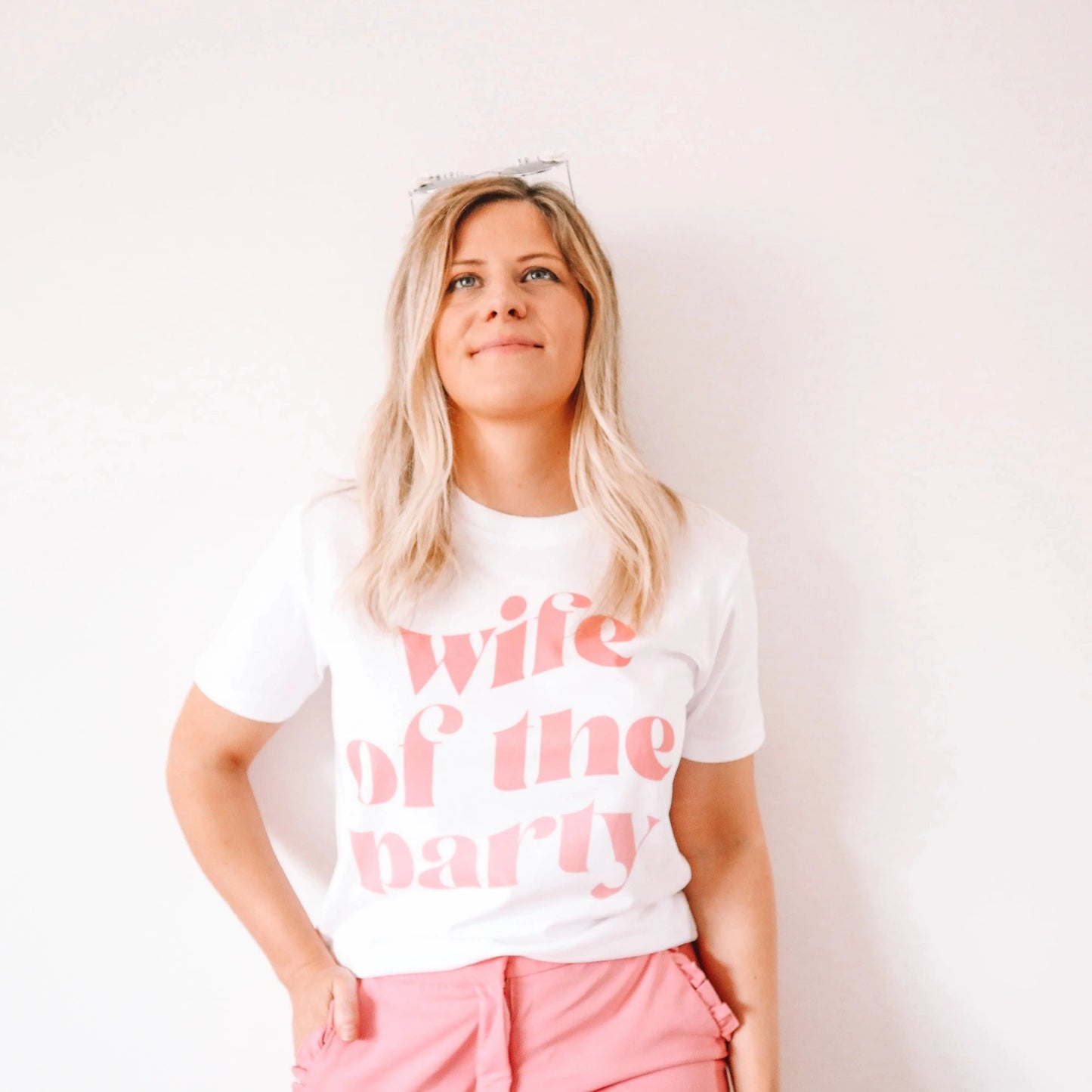 Woman wearing a white “wife of the party” hen party t-shirt with soft pink lettering and pink trousers, standing against a plain light background.