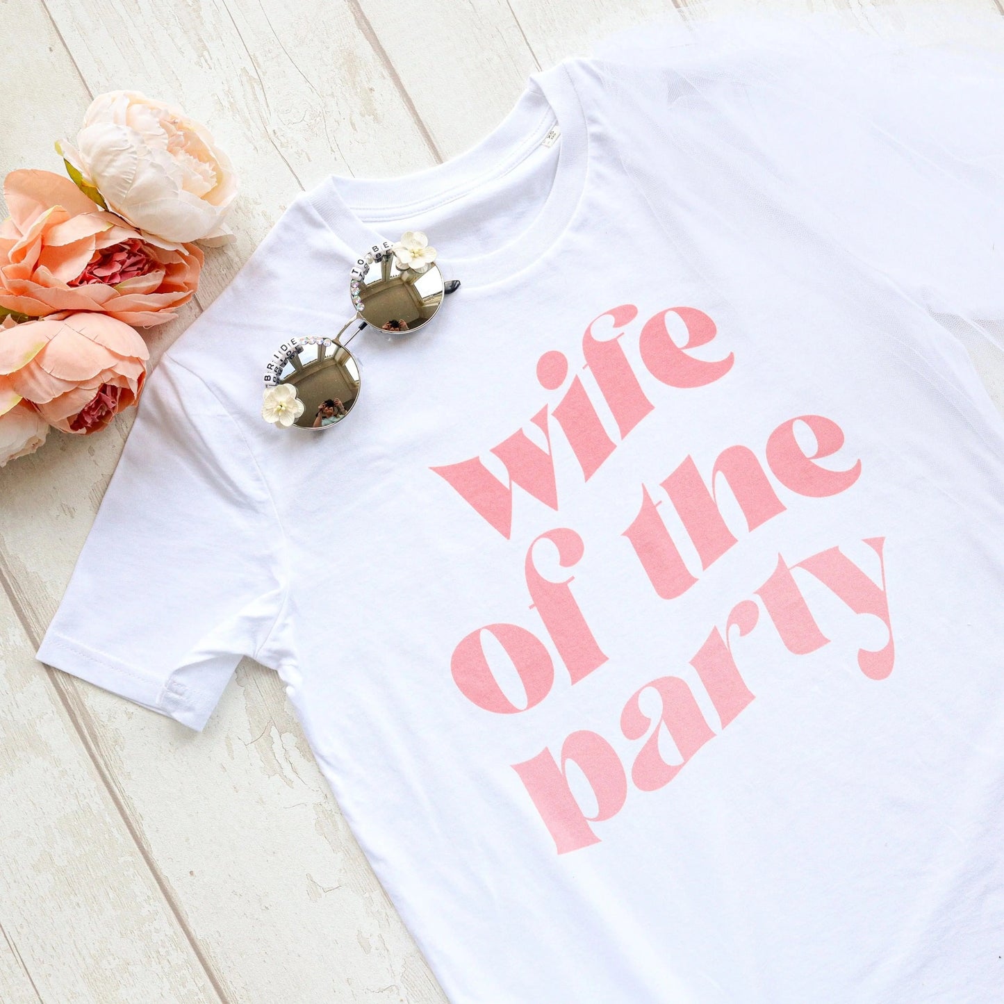 Styled flat lay of a white “wife of the party” hen party t-shirt with soft pink lettering, paired with bride sunglasses, a veil, and blush flowers on a light wooden background.