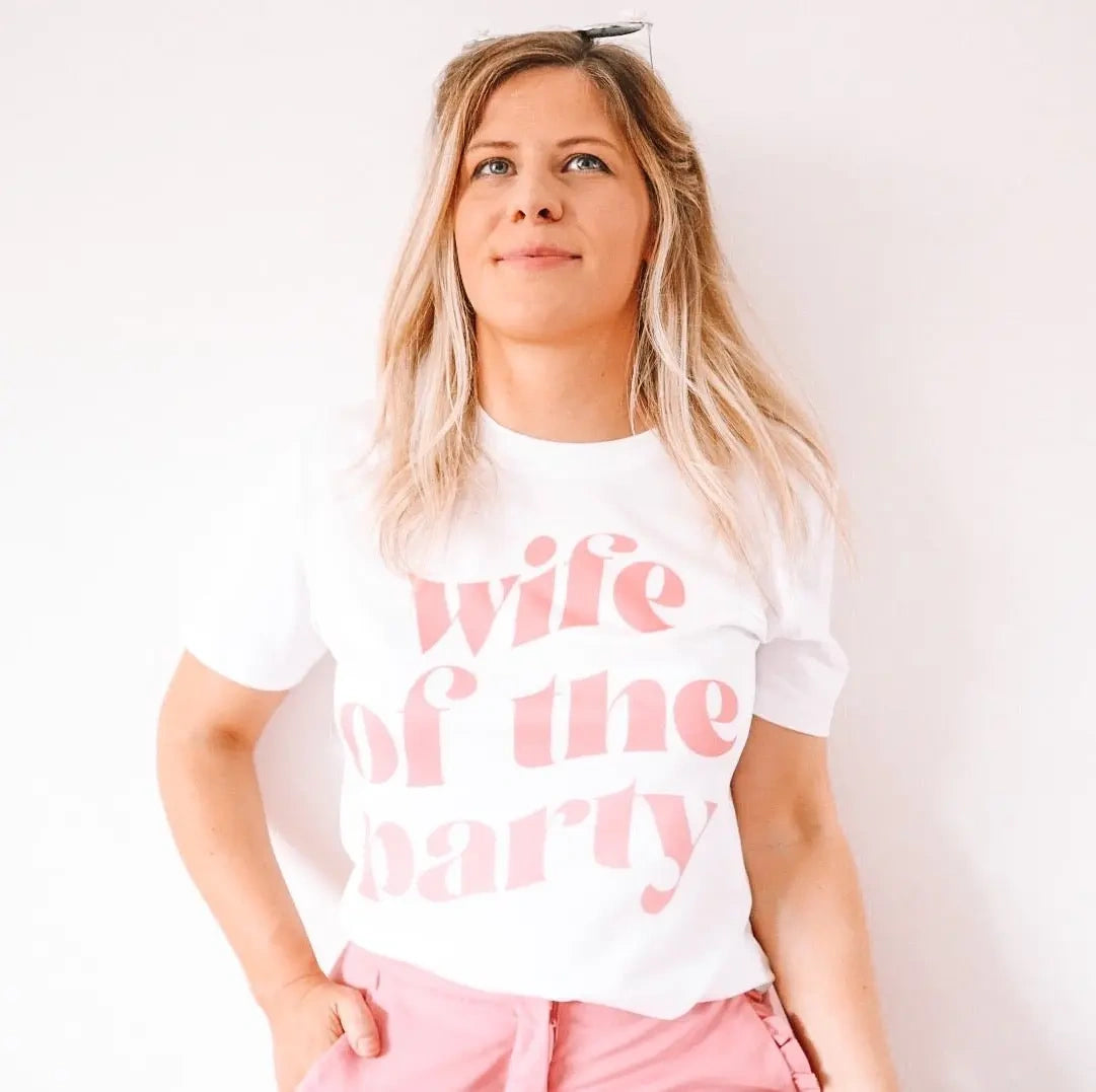 Woman wearing a white “wife of the party” hen party t-shirt with pale pink lettering, styled with pink bottoms against a soft neutral background.