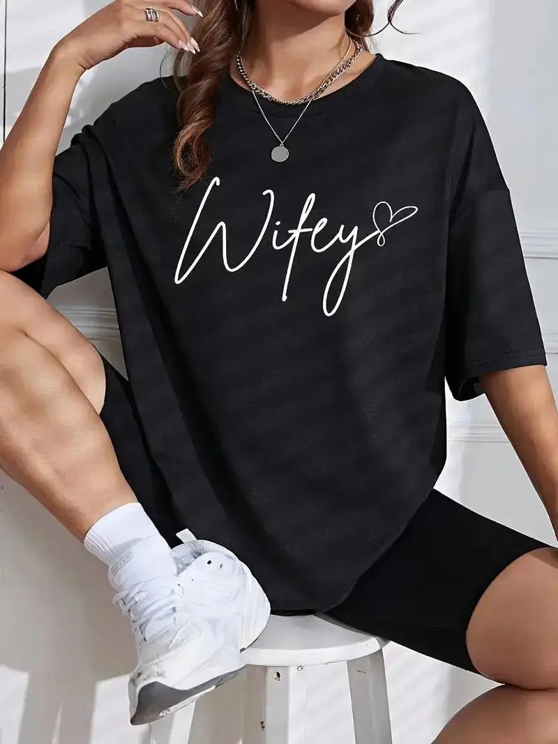 Person wearing a black t-shirt with 'Wifey' printed on it, sitting on a white stool.