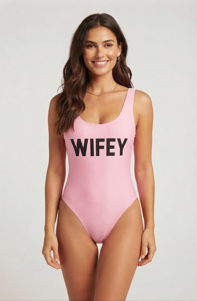 A woman modeling a light pink one-piece swimsuit with the word 'WIFEY' printed in bold black letters across the chest, set against a plain light gray studio background.