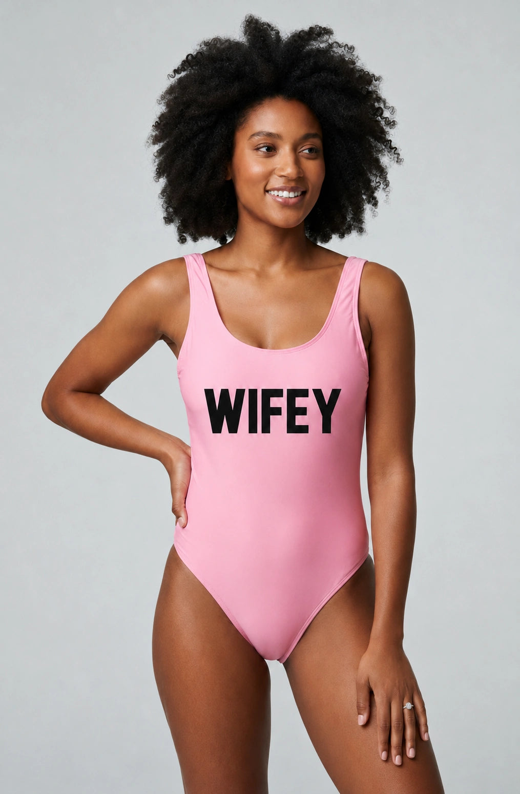 A woman modeling a pink one-piece swimsuit with the word 'WIFEY' printed in bold black letters across the chest, photographed against a light gray studio background.