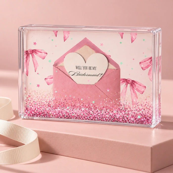 A clear acrylic keepsake box filled with pink glitter, featuring a printed pink invitation card with an envelope, heart, and bachelorette-themed graphics, displayed on a pink surface with a cream ribbon.