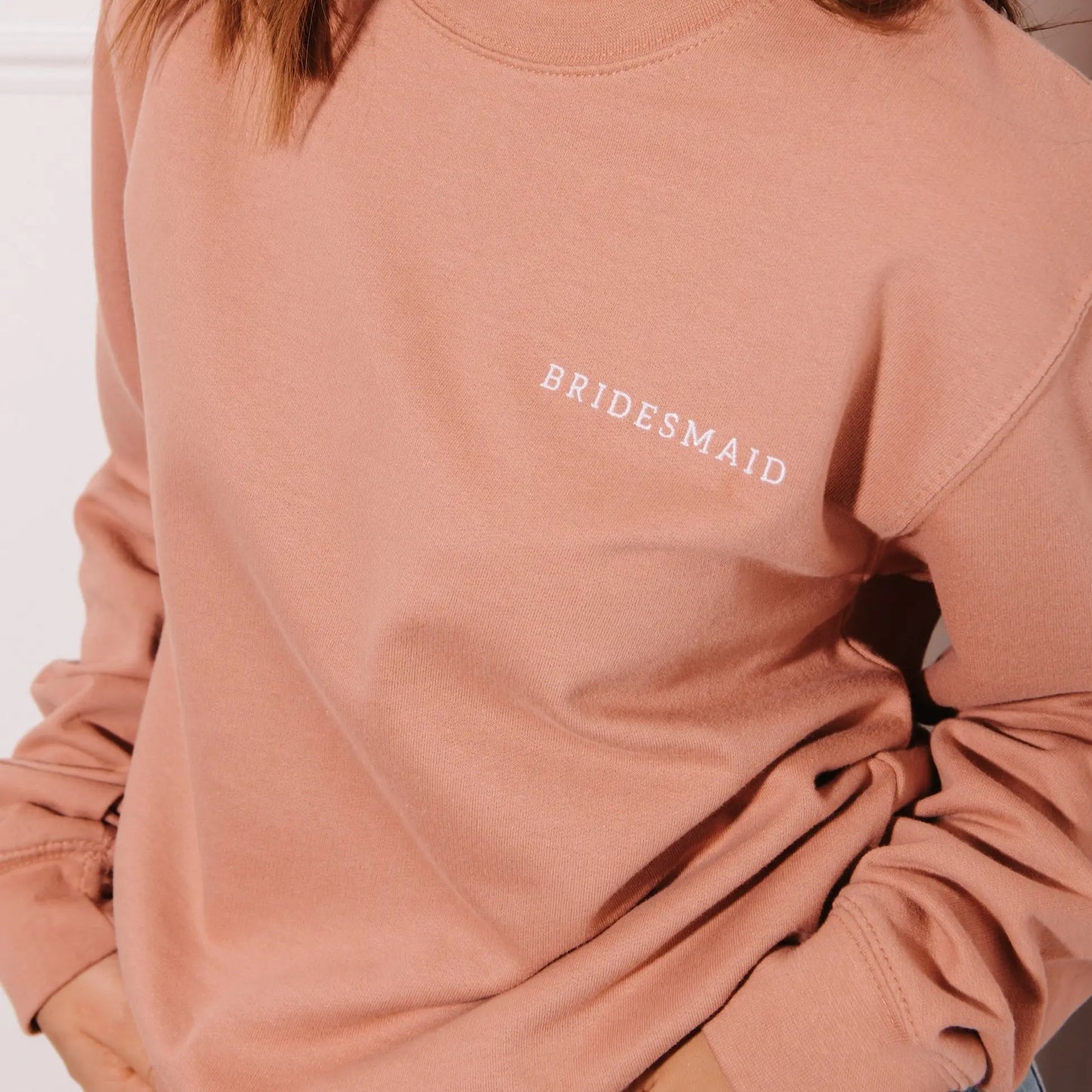 A close-up of a person wearing a dusty rose crewneck sweatshirt with "BRIDESMAID" embroidered in white serif capital letters on the left chest. The person has their hands in their pockets, and only their torso and a few strands of brown hair are visible against a neutral background.